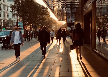 City street scene at sunset with people walking on a sidewalk lined with trees and buildings. The setting sun casts long shadows, and a double-decker bus is visible in the background.